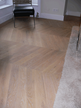 Holzboden im Fischgrätmuster mit einem Stuhl im Hintergrund.