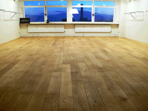 Helle, leere Raum mit Holzfußboden und großen Fenstern im Hintergrund.